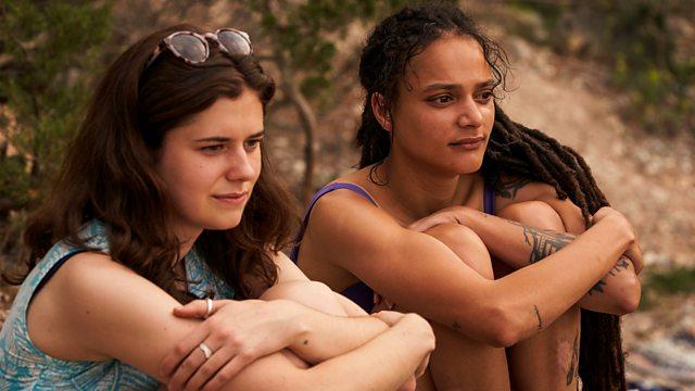 Two young women, both with long dark hair, one white and one black, are sitting in what seems to be a beach, looking at the horizon.