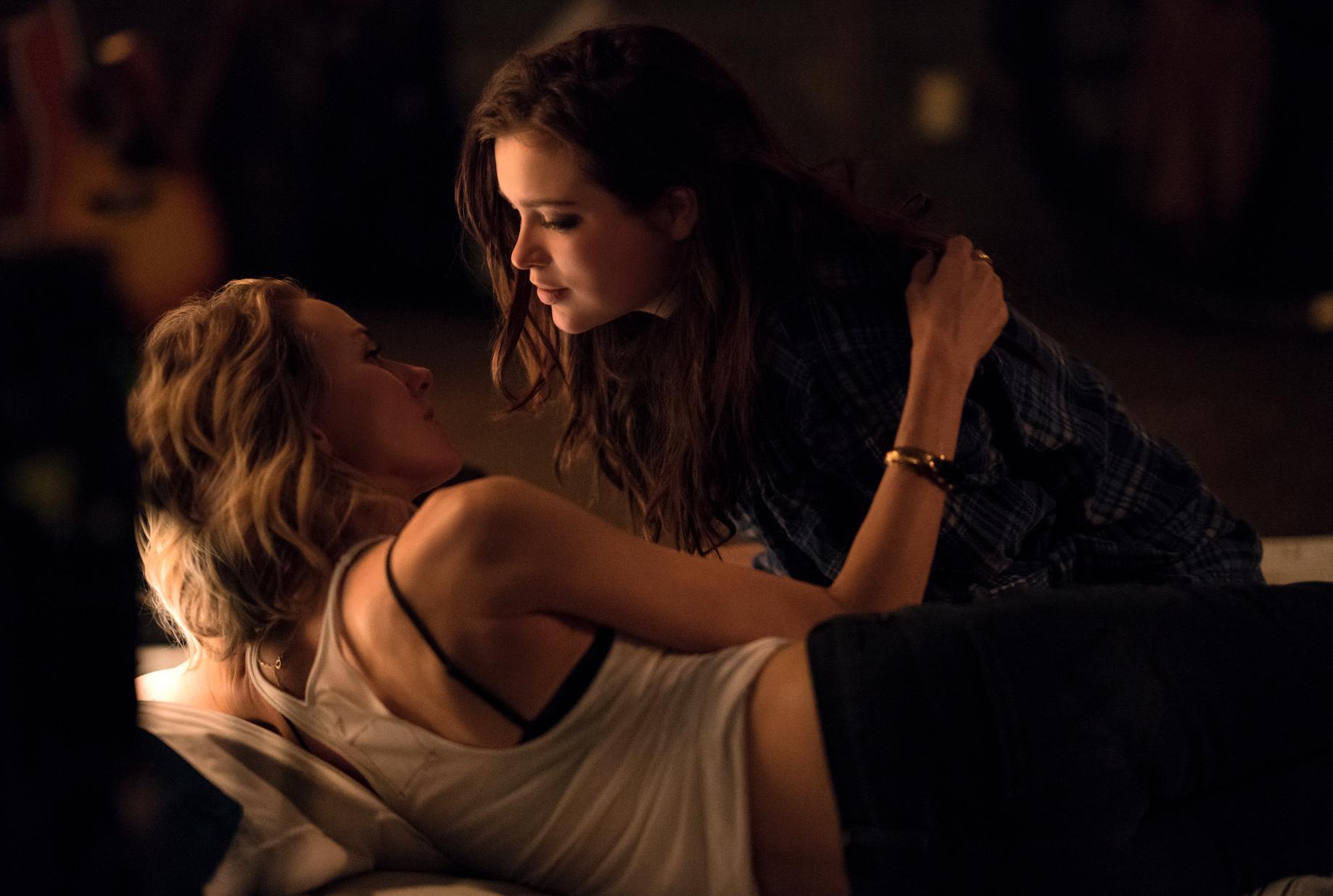 Two women are laying down, one of them is blonde and the other is a brunette. The light is subtle, intimate; they might be about to kiss.