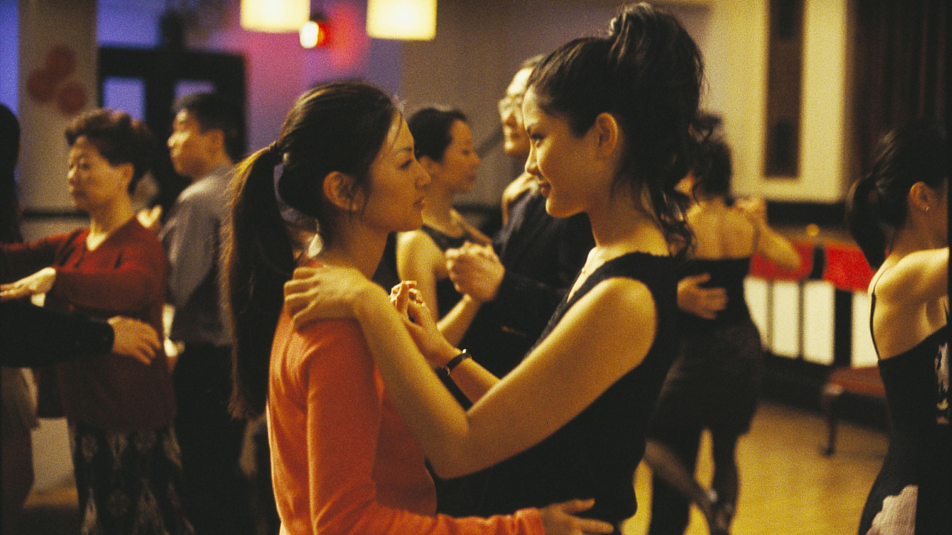 This image shows two women engaged in an intimate dance, holding each other closely and gazing into each other's eyes. The dimly lit, colorful environment suggests a social gathering or event.